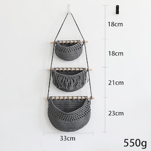 Puede incluir: Un colgante de pared de macram&eacute; gris con tres cestas tejidas. Las cestas est&aacute;n unidas a tacos de madera y suspendidas por una cuerda gris. La cesta inferior mide 33 cm de ancho y 23 cm de alto. El peso total es de 550 g.