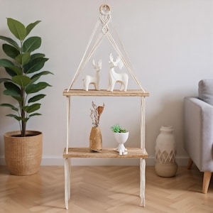 May include: A two-tier macrame hanging shelf with wooden shelves. The top shelf displays two white deer ornaments. The bottom shelf holds a twine-wrapped vase, a small white planter with a succulent, and dried flowers. The shelf is suspended by white macrame cords.