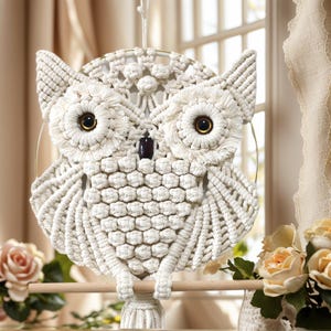 Könnte beinhalten: Handgefertigte Makramee-Eule zum Aufhängen. Die Eule besteht aus cremefarbenem Seil mit detaillierten Flügeln, Augen und einem Schnabel. Sie hängt an einer Holzstange. Der Hintergrund zeigt ein Fenster und Blumen.