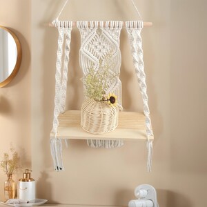 May include: A macrame wall shelf with a natural wood shelf and a woven basket vase holding dried flowers and a small sunflower. The shelf is suspended by a wooden dowel and white macrame cords.