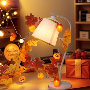 May include: A white desk lamp with a rectangular lampshade, adorned with autumn leaves, pumpkins, and acorns. The lamp is surrounded by fairy lights, creating a warm, autumnal atmosphere. A gift box is also visible.