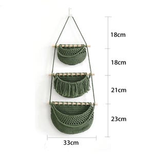 Puede incluir: Un colgador de plantas de macram&eacute; de tres niveles en verde oliva. Cada nivel es una cesta tejida suspendida por una cuerda y una clavija de madera. La cesta inferior mide 33 cm de ancho y 23 cm de alto. Las dos cestas superiores miden 18 cm de alto. La cesta del medio tiene flecos.