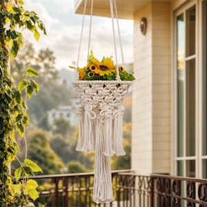 Könnte beinhalten: Ein cremefarbener Makramee-Pflanzenhalter mit korbförmiger Struktur, gefüllt mit gelben Sonnenblumen. Der Halter wird an einem weißen Seil aufgehängt. Das Makramee hat ein gewebtes Design mit langen Fransen. Auf einem Balkon.