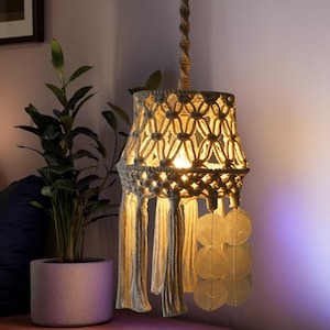 Puede incluir: Lámpara colgante de macramé hecha a mano con una pantalla de color crema y borlas. La pantalla tiene un patrón geométrico y está suspendida de una cuerda trenzada. Acentos decorativos de concha cuelgan debajo.
