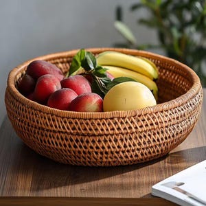Puede incluir: Una cesta redonda de mimbre tejida llena de fruta fresca. La cesta contiene melocotones rojos, plátanos amarillos y un mango amarillo. La cesta está sobre una superficie de madera, lo que sugiere una cocina o comedor.
