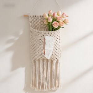 Op de afbeelding: Een crèmekleurige macramé wandzak met een houten deuvel en franjes. Het bevat een boeket roze tulpen en een tissue. Het geweven ontwerp voegt een bohemien touch toe aan de interieurdecoratie.