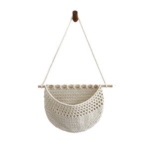 Puede incluir: Una cesta colgante de macram&eacute; de color crema, con una clavija de madera y una clavija de madera marr&oacute;n oscuro para colgarla. La cesta tiene forma semicircular con un dise&ntilde;o tejido. Se suspende con una cuerda de color crema.