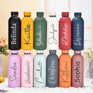 May include: A collection of personalized water bottles in various colors, including black, yellow, green, white, red, and blue. Each bottle features a different name in a stylish script font. The bottles are cylindrical with a screw-on cap.