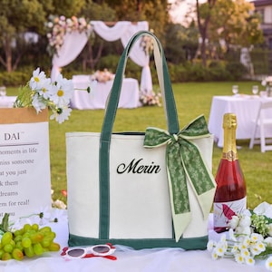 May include: A cream-colored canvas tote bag with dark green trim and handles. The front features the name Merin embroidered in black script. A decorative green ribbon with a subtle floral pattern is tied to one of the handles in a bow.