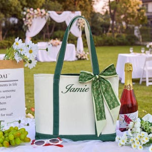 Puede incluir: Un bolso tote de lona color crema con bordes y asas de color verde oscuro. El nombre Jamie está bordado en cursiva verde oscuro en la parte delantera. Una cinta decorativa con estampado verde está atada en un lazo en el asa derecha.