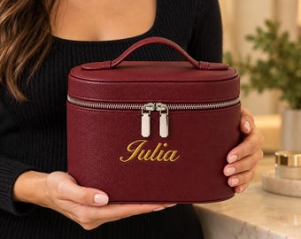 May include: A burgundy oval-shaped cosmetic case with a textured finish, a top handle, and a silver-toned double zipper closure. The name Julia is embroidered in gold script on the front center of the case.