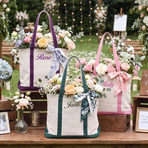 May include: Three off-white canvas tote bags with colored trim and handles in purple, teal, and pink. Each bag is embroidered with a name: Rose, Jane, or Molly. The bags are filled with assorted pastel flowers and tied with patterned fabric ribbons.