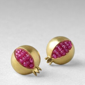May include: A pair of gold earrings shaped like pomegranates, each with a half-circle cut out to reveal a cluster of pink gemstones.