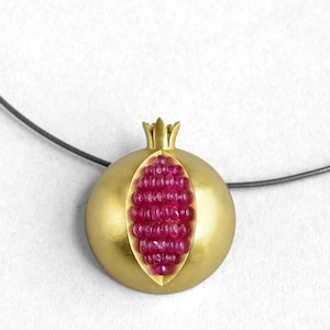 Pu&ograve; includere: Una collana d'oro con un ciondolo a forma di melograno. Il ciondolo &egrave; aperto per rivelare un gruppo di pietre di rubino rosse.