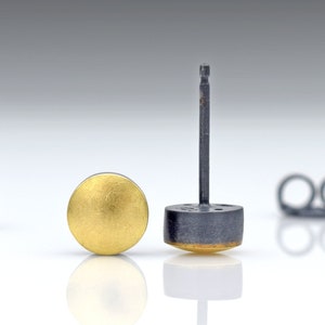 May include: A pair of gold and black stud earrings. The earrings are round and have a smooth finish. The earrings are on a white surface.