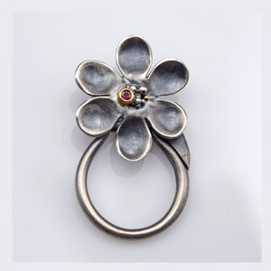 Puede incluir: Un anillo de plata con un diseño de flor. La flor tiene seis pétalos y una pequeña piedra preciosa roja en el centro. El anillo está oxidado y tiene un aspecto rústico.