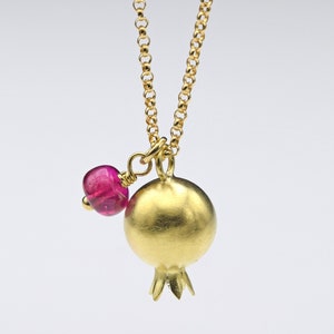 Può includere: Una collana dorata con un ciondolo a forma di melograno e una piccola gemma rosa. Il ciondolo a melograno ha una superficie strutturata e un gambo dettagliato. Anche la catena della collana è dorata.