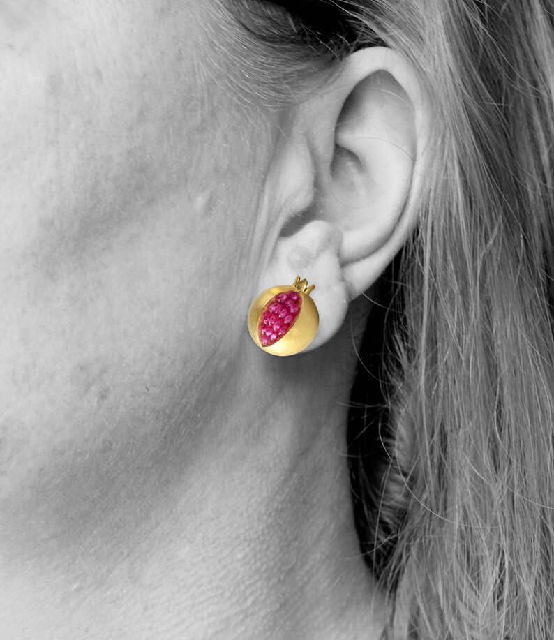 Pu&ograve; includere: Un paio di orecchini d'oro a forma di melograno con pietre di rubino rosse.