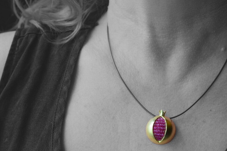 Pu&ograve; includere: Una collana d'oro con un ciondolo a forma di melograno. Il ciondolo &egrave; in oro e ha un centro di rubino rosso.