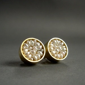 May include: A pair of gold-colored stud earrings, each featuring a cluster of small, white pearls. The earrings have a circular design, with the pearls filling the interior of the gold setting. The background is a solid, dark gray.