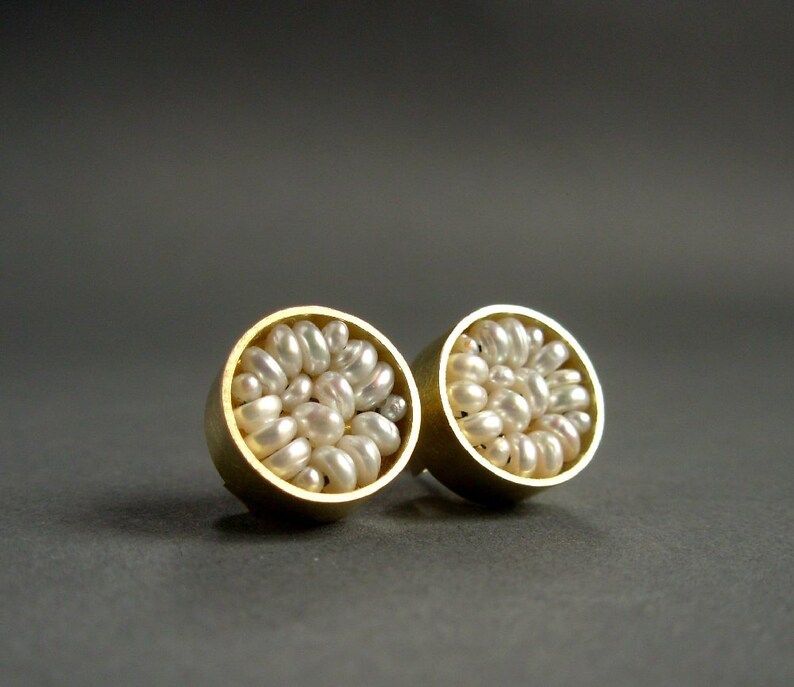 May include: A pair of gold-colored stud earrings, each featuring a cluster of small, white pearls. The earrings have a circular design, with the pearls filling the interior of the gold setting. The background is a solid, dark gray.