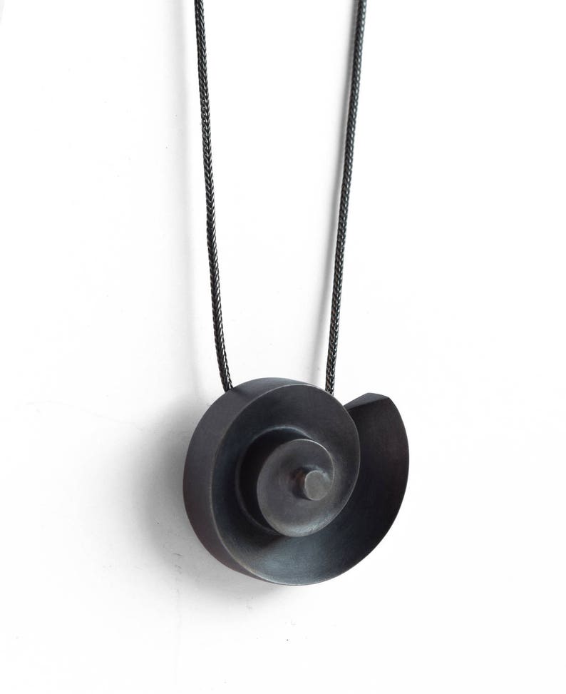 Pu&ograve; includere: Una collana con un ciondolo in argento ossidato nero a forma di spirale. Il ciondolo &egrave; sospeso da un cordoncino nero.