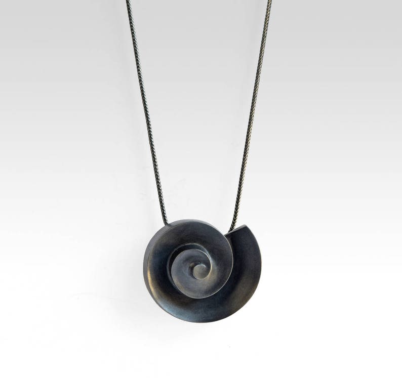 Pu&ograve; includere: Una collana d'argento con un ciondolo nero a forma di spirale. Il ciondolo &egrave; una rappresentazione stilizzata di una conchiglia.