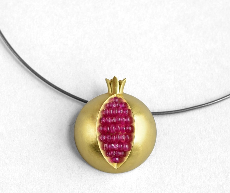 Pu&ograve; includere: Una collana d'oro con un ciondolo a forma di melograno. Il ciondolo &egrave; aperto per rivelare un gruppo di pietre di rubino rosse.