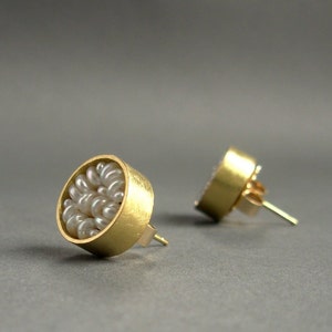 May include: A pair of gold-colored stud earrings. Each earring features a circular design filled with small, white pearls. The earrings have a post and backing for secure wear, set against a gray background.