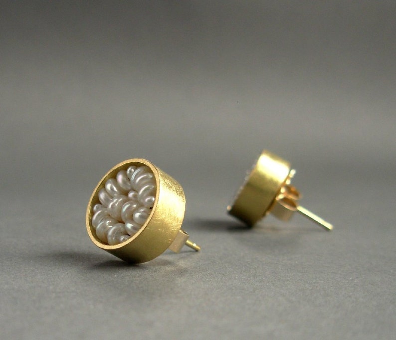 May include: A pair of gold-colored stud earrings. Each earring features a circular design filled with small, white pearls. The earrings have a post and backing for secure wear, set against a gray background.