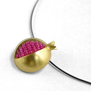 Pu&ograve; includere: Una collana con un ciondolo d'oro a forma di melograno. Il melograno &egrave; pieno di pietre preziose rosa.