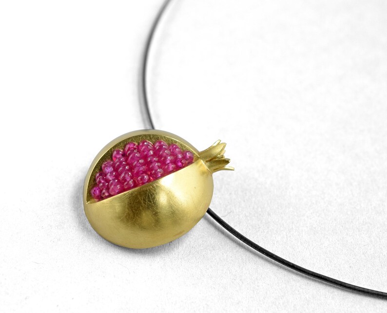 Pu&ograve; includere: Una collana con un ciondolo d'oro a forma di melograno. Il melograno &egrave; pieno di pietre preziose rosa.