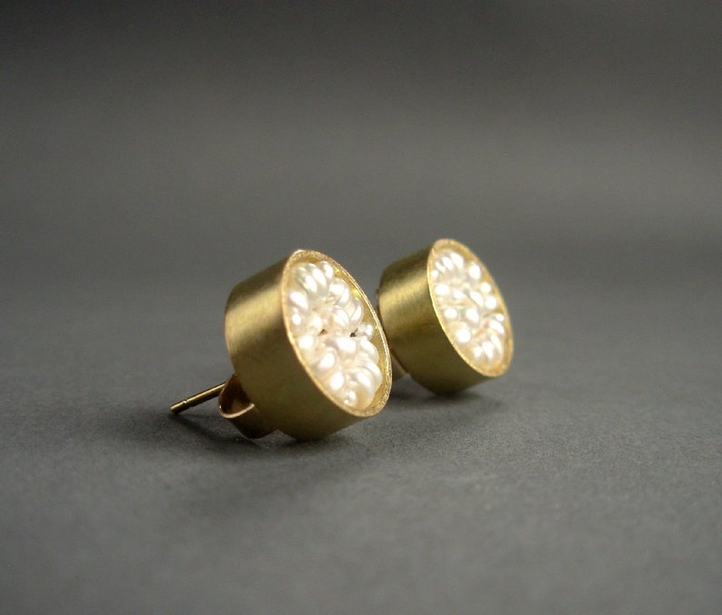 May include: A pair of gold-colored stud earrings. Each earring features a circular design filled with small, white pearls. The earrings have a post and backing for secure wear. The background is a solid, dark gray.