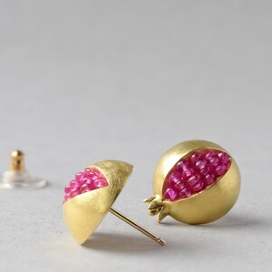 Pu&ograve; includere: Un paio di orecchini d'oro a forma di melograno, con pietre preziose rosa all'interno.