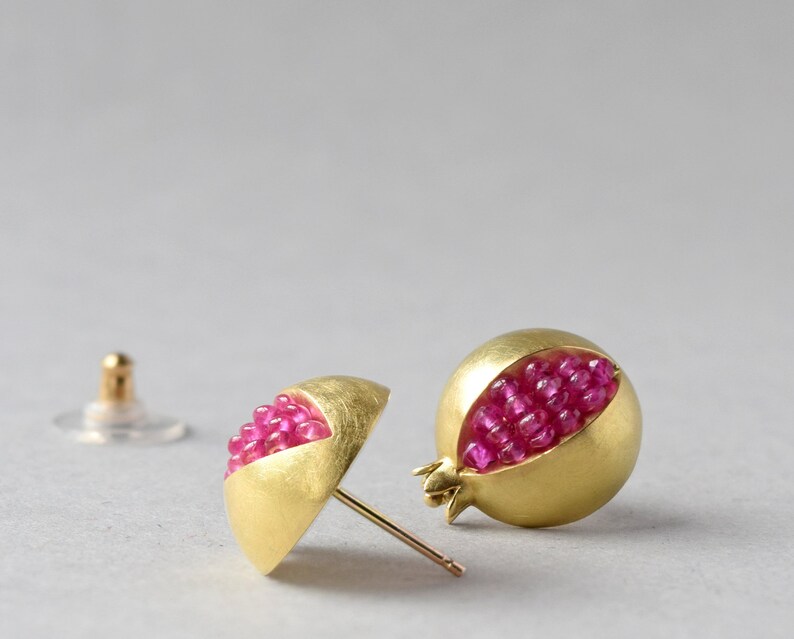 Pu&ograve; includere: Un paio di orecchini d'oro a forma di melograno, con pietre preziose rosa all'interno.