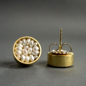 May include: A pair of gold-tone stud earrings. Each earring features a circular design filled with small, irregularly shaped white pearls. The earrings have a post and butterfly backing for secure wear. The background is a solid gray.