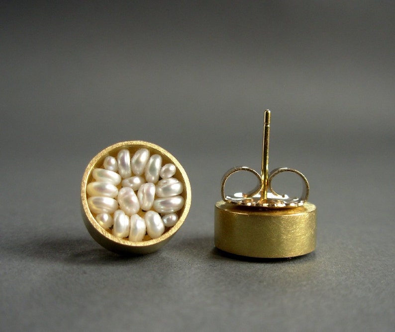 May include: A pair of gold-tone stud earrings. Each earring features a circular design filled with small, irregularly shaped white pearls. The earrings have a post and butterfly backing for secure wear. The background is a solid gray.