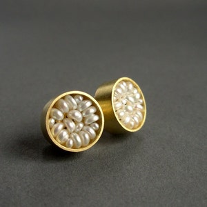 May include: A pair of gold-colored stud earrings. Each earring features a circular design filled with small, white, irregularly shaped pearls. The earrings are set against a dark gray background, highlighting their metallic sheen and pearl texture.