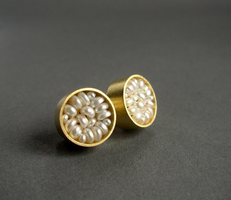 May include: A pair of gold-colored stud earrings. Each earring features a circular design filled with small, white, irregularly shaped pearls. The earrings are set against a dark gray background, highlighting their metallic sheen and pearl texture.