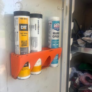 Pu&ograve; includere: Un supporto in plastica arancione montato all'interno di un armadio contiene tre tubi di grasso. I tubi sono etichettati "CAT Utility Grease", "CATERPILLAR" e "LUCAS OIL PRODUCTS MARINE GREASE". L'armadio &egrave; bianco.