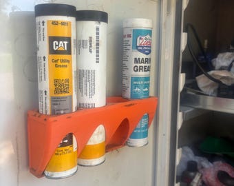 Puede incluir: Un soporte de plástico naranja montado dentro de un armario contiene tres tubos de grasa. Los tubos están etiquetados como "CAT Utility Grease", "CATERPILLAR" y "LUCAS OIL PRODUCTS MARINE GREASE". El armario es blanco.