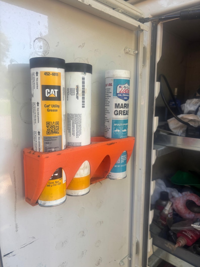 Pu&ograve; includere: Un supporto in plastica arancione montato all'interno di un armadio contiene tre tubi di grasso. I tubi sono etichettati "CAT Utility Grease", "CATERPILLAR" e "LUCAS OIL PRODUCTS MARINE GREASE". L'armadio &egrave; bianco.