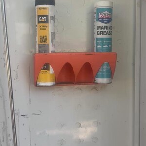 Pu&ograve; includere: Un portapacchi in plastica arancione montato su una porta in metallo bianco contiene tre contenitori cilindrici. I contenitori includono CAT Utility Grease e Lucas Oil Products Marine Grease. La porta ha un chiavistello e altri accessori.