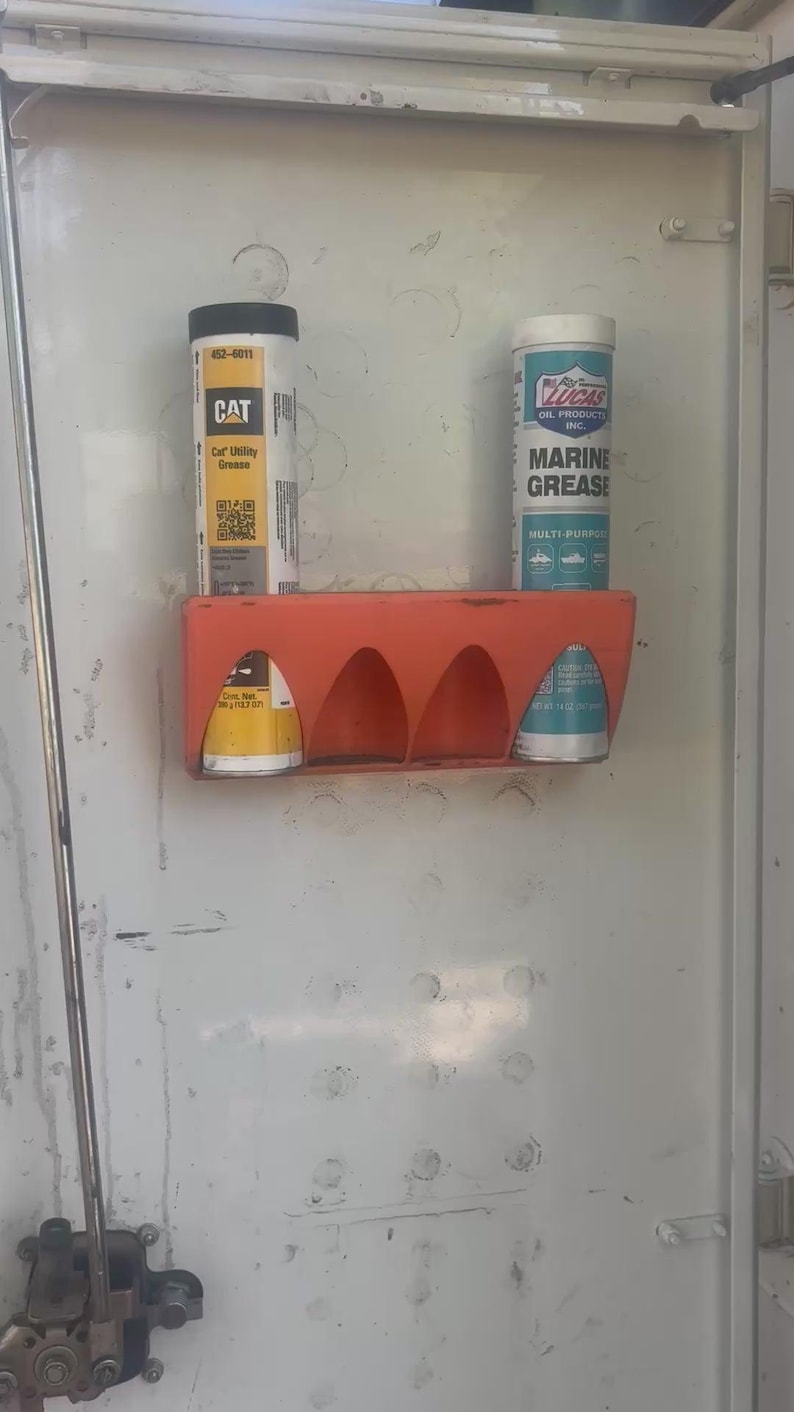Pu&ograve; includere: Un portapacchi in plastica arancione montato su una porta in metallo bianco contiene tre contenitori cilindrici. I contenitori includono CAT Utility Grease e Lucas Oil Products Marine Grease. La porta ha un chiavistello e altri accessori.