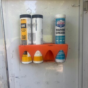Pu&ograve; includere: Un supporto arancione che contiene vari contenitori cilindrici. I contenitori includono "CAT Car Utility Grease" e "Lucas Marine Grease". Il supporto &egrave; montato su una superficie bianca.