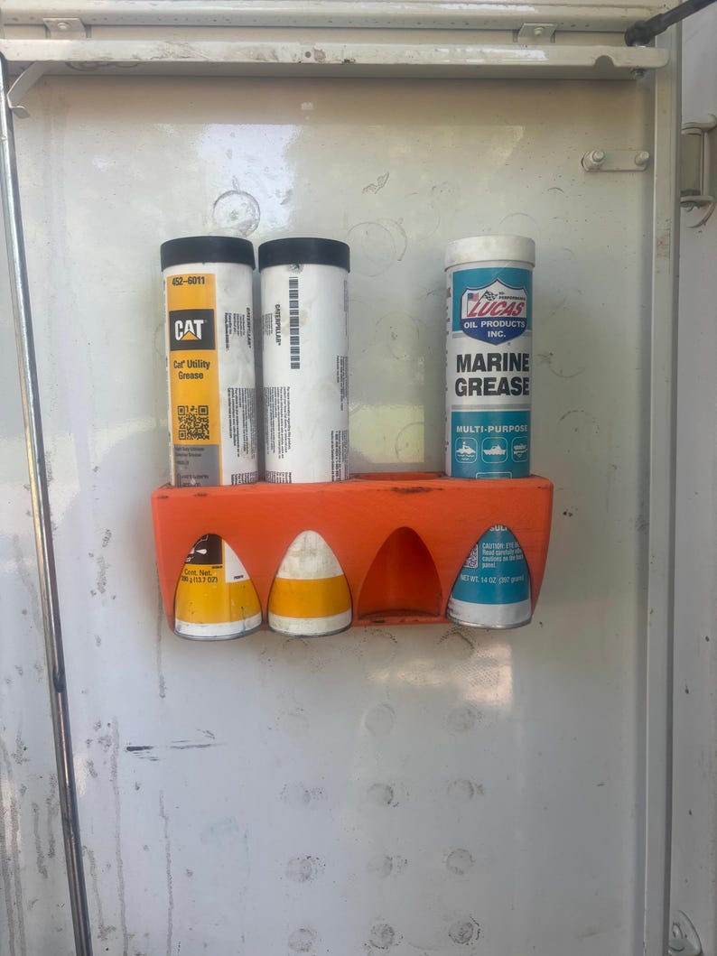 Pu&ograve; includere: Un supporto arancione che contiene vari contenitori cilindrici. I contenitori includono "CAT Car Utility Grease" e "Lucas Marine Grease". Il supporto &egrave; montato su una superficie bianca.