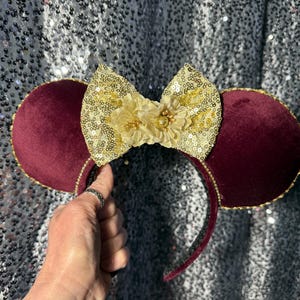 May include: Burgundy velvet mouse ears with gold trim and a gold sequined bow. The bow features a gold flower in the center. The headband is black with a burgundy velvet lining.