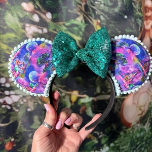 May include: A black headband with two round ears featuring a colorful design with a castle and planets. The ears are trimmed with white pom-poms. A large, sequined emerald green bow sits between the ears.