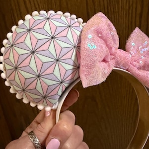 May include: Headband featuring two circular ears and a central bow. The ears are covered in a fabric with a geometric pattern of triangles in pastel pink, purple, and mint green, finished with a white pom-pom trim. The bow is covered in pink sequins.
