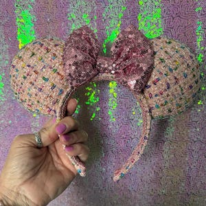 May include: Pink tweed mouse ears headband with a sequined bow. The ears are embellished with colorful flecks, and the bow is covered in pink sequins. The headband is held by a hand with pink nail polish.